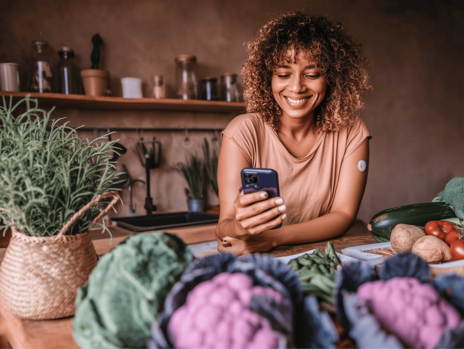 Smiling woman in a kitchen wearing a continuous glucose monitor on her arm, looking at her smartphone, with fresh vegetables and herbs on the counter in front of her.