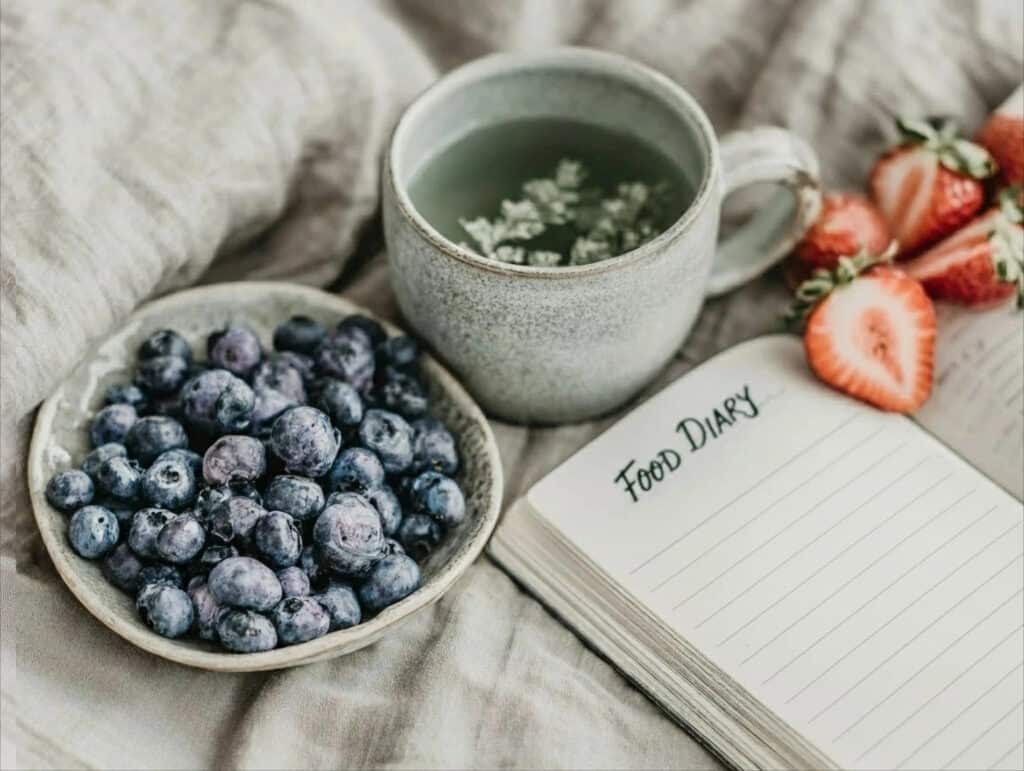 Open food diary on a bed with fresh blueberries, sliced strawberries, and a cup of herbal tea, symbolising mindful eating and tracking how food affects the body.