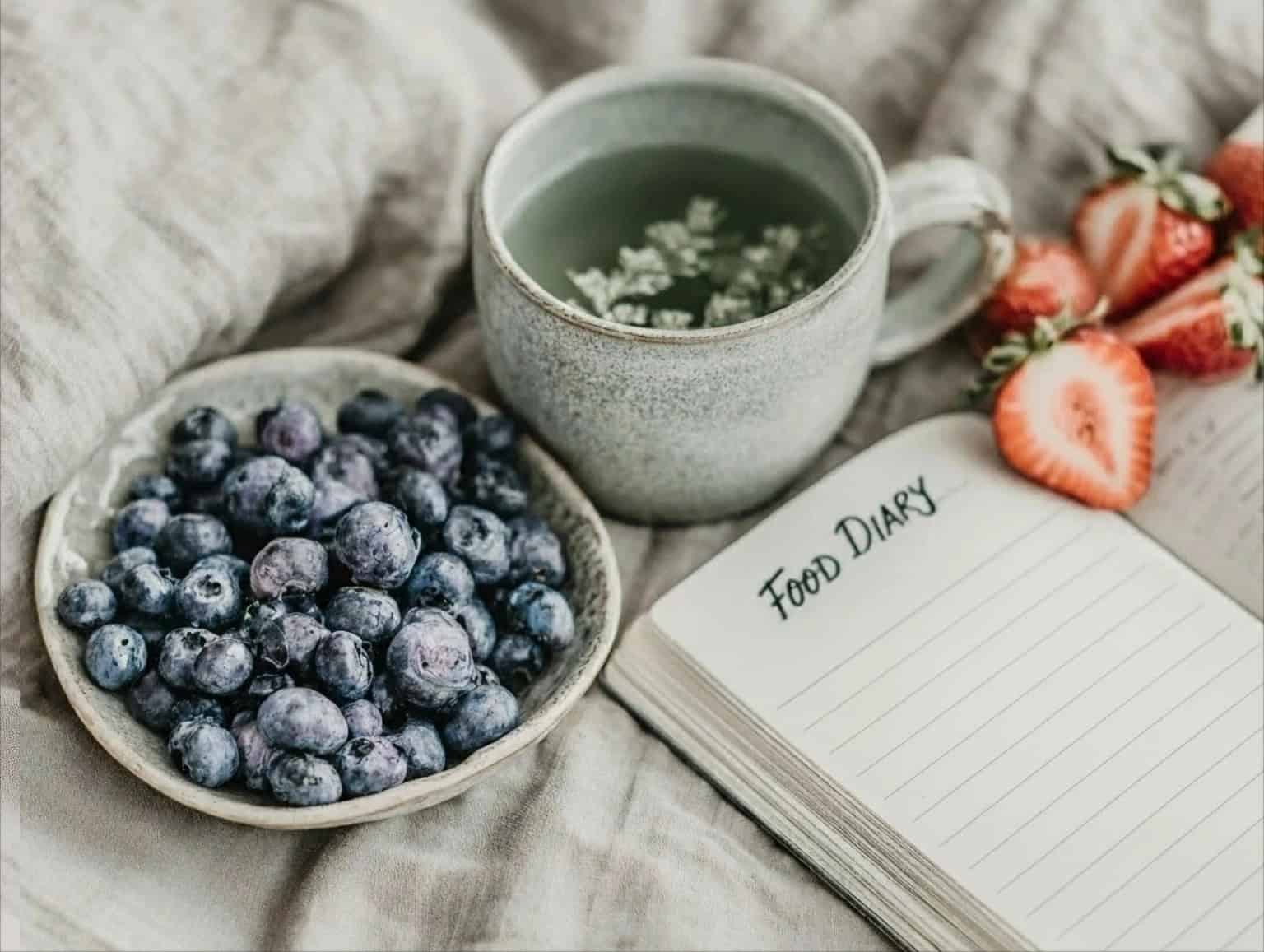 Open food diary on a bed with fresh blueberries, sliced strawberries, and a cup of herbal tea, symbolising mindful eating and tracking how food affects the body.